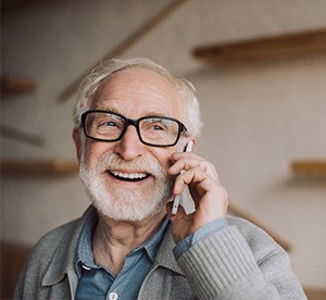 Man with white hair and beard in gray sweater and black glasses talking on his cell phone