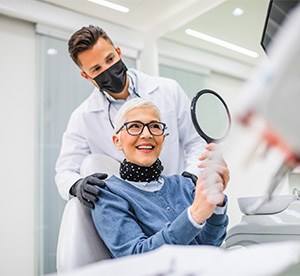 Woman in blue shirt looking at hand mirror with dentist standing behind her