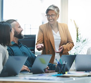 Woman with gray hair in brown jacket presenting to colleagues around a table